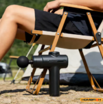 Person using a portable percussion massage gun outdoors while relaxing on a camping chair, demonstrating its compact and travel-friendly design – UK